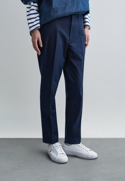 Navy blue pants with a tailored fit, featuring a smooth texture and subtle crease detailing, paired with white sneakers. Striped shirt visible.