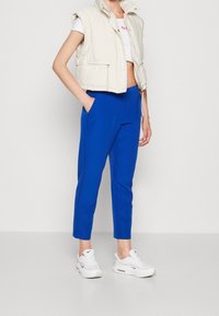 Personne portant un pantalon bleu fuselé, un crop top blanc, un gilet matelassé écru et des baskets blanches Nike, debout avec une main dans la poche.