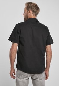 Svart kortermet skjorte med en myk tekstur, knappekrage og rett kant, båret over lyse grå denimjeans.
