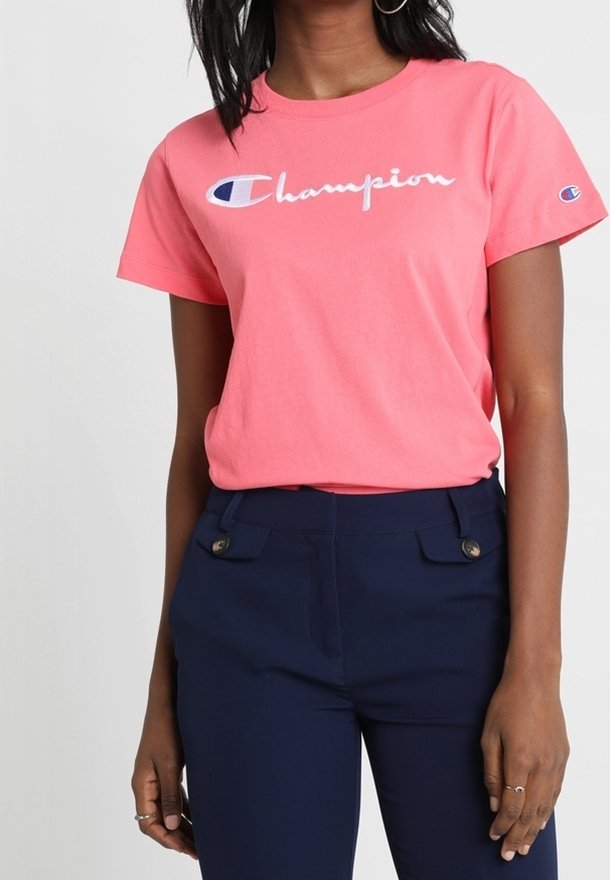 Vaaleanpunainen puuvillapaita, jossa on valkoinen 'Champion'-logo edessä. Yhdistetty tummansiniset slim-fit-housut, joissa on nappikoristeet.