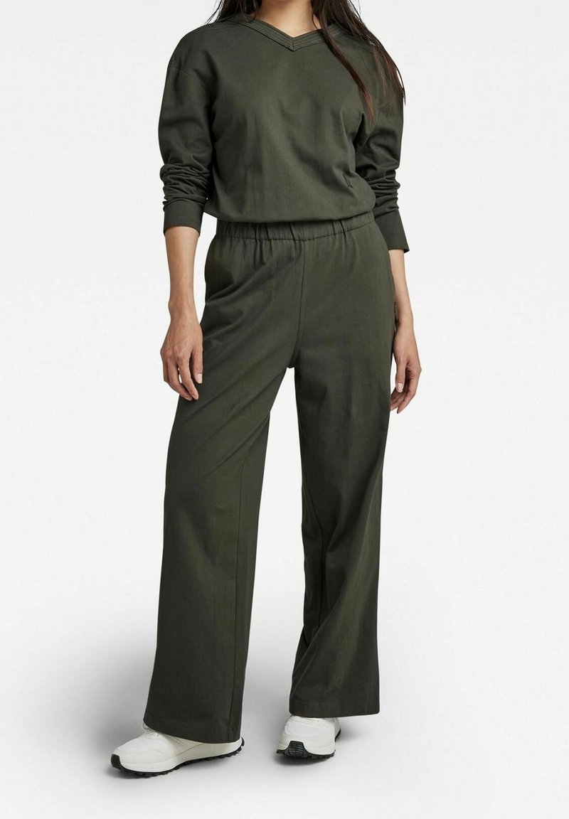 Femme portant un haut à manches longues vert foncé et un pantalon large avec des baskets blanches, debout devant un fond clair uni.