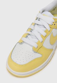 Kollane ja valge tennisekets nahast ja võrgust materjalidest, ümmargune varvas, aukudega detailid, valged paelad ja Nike swooshi logo.