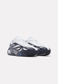 Ζευγάρι αθλητικών παπουτσιών, λευκά και μαύρα, με μπλε λογότυπα, κυματιστό σχεδιασμό και υφασμένες λαστιχένιες σόλες σε απλό φόντο.