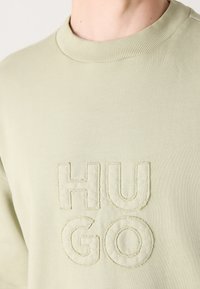 Close-up van een lichtbeige sweatshirt met verhoogde gestikte letters die "HUGO" spellen op de borst en een geribbelde ronde halskraag.
