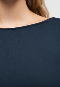 Κοντινό πλάνο του λαιμού και του ώμου ενός ατόμου που φοράει μπλε μπλουζάκι με στρογγυλή λαιμόκοψη, με ορατά μακριά ξανθά μαλλιά.