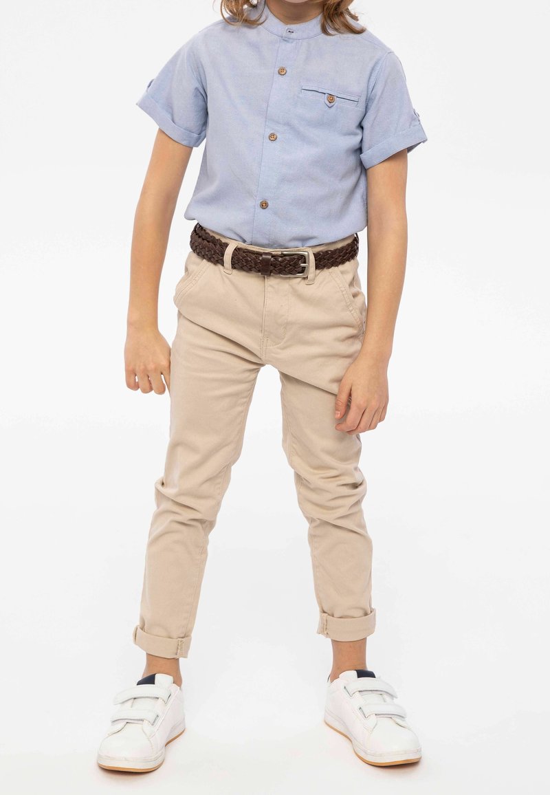 Enfant portant une chemise boutonnée à manches courtes bleu clair, un pantalon beige retroussé avec une ceinture marron tressée et des baskets blanches.