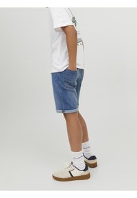 Pantalones cortos de mezclilla, de color azul claro con dobladillos enrollados, combinados con zapatillas blancas y calcetines de canalé. Mano del modelo en los bolsillos, vista lateral.