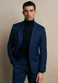 Granatowy garnitur w kratę, zestawiony z czarnym golfem. Garnitur ma dopasowany krój i wyróżnia się pojedynczym guzikiem oraz bocznymi kieszeniami.