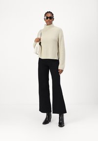 Beige ribbet rullekravegenser med bred silhuett, sammen med svarte utvidede bukser og svarte høyhælede støvler; modellen bruker solbriller.
