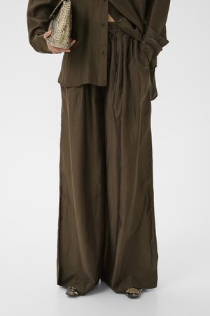 Luźne ciemnooliwkowe spodnie zestawione z pasującą koszulą, trzymająca beżową torebkę ze skóry węża i nosząca buty w wzór skóry węża.