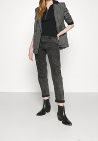 Blazer grigio sopra un top nero a coste, abbinato a jeans neri scoloriti. Il modello indossa stivaletti neri con tacco a blocco. Design semplice e strutturato.