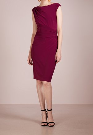 Femme portant une robe prune sans manches, longueur genou, ajustée avec un détail froncé sur le côté, et des talons noirs à bride cheville, se tenant devant un fond beige.