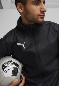 Schwarze Zip-Jacke mit hohem Kragen, versehen mit einem weißen Puma-Logo. Die Person hält einen silbernen und schwarzen Fußball mit ähnlichem Branding.