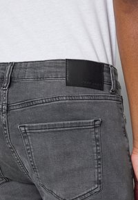 Vaqueros de mezclilla gris con un acabado texturizado, que cuentan con un parche de cuero negro en la cintura y detalles de costuras visibles. Dos bolsillos traseros.