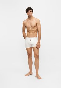Jeune homme à la silhouette sportive, debout pieds nus, portant un boxer blanc sur un fond blanc uni.