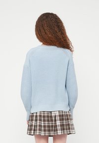 Pull bleu clair en tricot avec un col rond et des poignets côtelés, associé à une jupe plissée à carreaux marron. Vue de dos montrant la texture et le design.