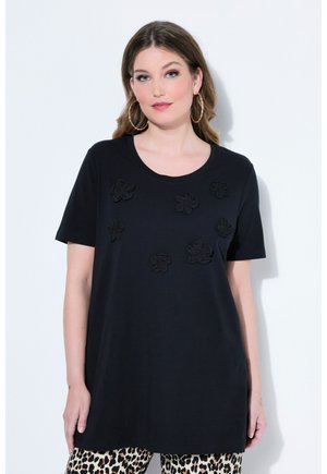 Kvinde med lyst brunt hår iført sort top med korte ærmer og blomsterapplikationer samt leopardmønstrede bukser, poserer mod en hvid væg.