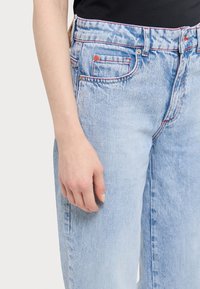 Jeans in denim blu chiaro con vestibilità comoda, dettagli in cucitura rossa, tasche anteriori e chiusura con bottone. La texture appare leggermente sbiadita.