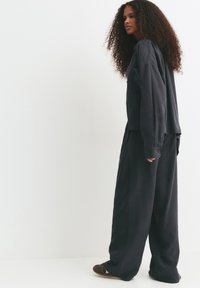 Woman with long curly hair wearing loose black shirt, wide black pants, and brown sneakers standing against a white background.