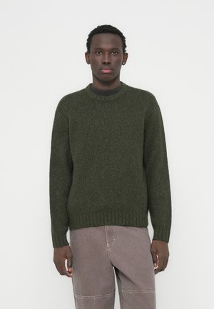 Pull tricot vert foncé avec un col rond et un ourlet côtelé, associé à un pantalon gris clair. La texture est épaisse et chaude.