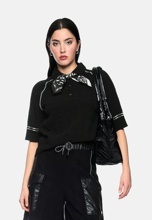 Young woman with long dark hair wears black knit top, cargo pants with leather pockets, a patterned neck scarf, and carries a black shiny bag.