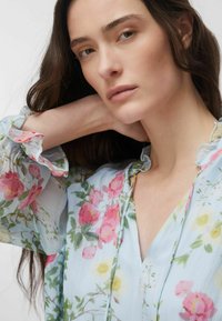 Gros plan sur une femme aux longs cheveux bruns portant un chemisier bleu clair avec des motifs floraux roses, jaunes et verts, sa main reposant derrière son cou.