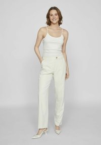 Top bianco, pantaloni beige chiaro su misura con chiusura a bottone e mules coordinate con tacco. Tessuto morbido e design a gamba dritta.