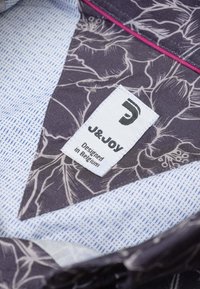 Grijze stof met bloemmotieven en een textuur van lichtblauwe voering. Etiket zegt "J&JOY, Ontworpen in België." Dikkere ontwerpelementen zichtbaar.