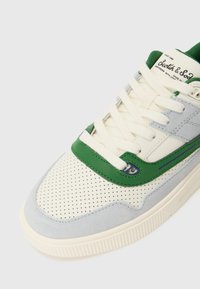 Sneaker med en blanding af grå ruskind og cremefarvet læder, grønne accenter, perforerede overflader og en beige gummisål med teksturerede detaljer.