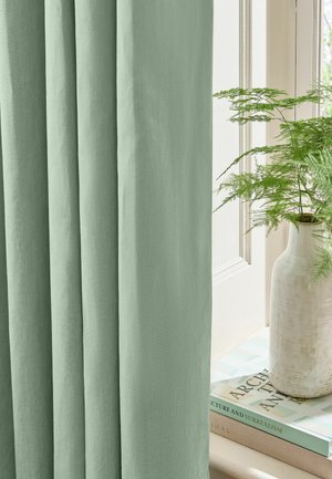 Tenda verde chiaro che copre parzialmente la finestra con un vaso bianco che contiene una felce verde sul davanzale accanto a libri impilati.