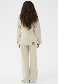 Beige, skræddersyet blazer med en subtil tekstur, parret med brede bukser. Har et kvadratisk mærke på bagsiden af jakken.