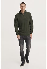 Donkergroen gebreid sweatshirt met een ritskraag, geribbelde textuur en een ontspannen pasvorm, gecombineerd met vervaagde zwarte jeans en zwarte sneakers.