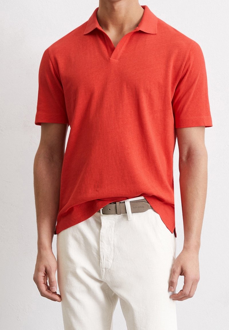 Homme portant un polo rouge à manches courtes légèrement rentré dans un pantalon blanc avec une ceinture grise, debout devant un fond uni.