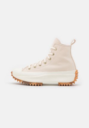 Beżowy trampek z płótna z wysoką cholewką, białą grubą podeszwą i brązowym, szorstkim bieżnikiem, metalowymi oczkami i pętelką do ściągania na pięcie.