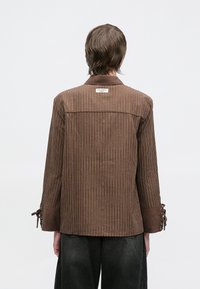 Ruda dryžuota marškinėliai su apykakle, zomšos rankovėmis ir raišteliais rankogaliuose. Etiketė matoma nugaroje, dėmesį patraukiantis tekstūruotas paviršius.