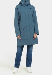 Vattenavvisande blå jacka med huva, dragkedja fram och fickor, långa ärmar och rak nederkant. Kombinerad med blå jeans och vita skor.