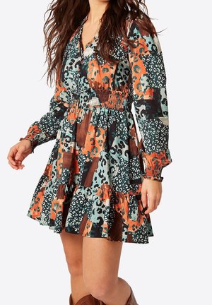 Femme portant une mini-robe à manches longues et col en V avec un imprimé animal abstrait orange, bleu sarcelle, noir et blanc et une taille froncée.