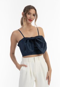 Námořnický saténový crop top s velkou mašlí na přední straně, spárovaný s bílými kalhotami s vysokým pasem. Viditelné tenké ramenní popruhy.