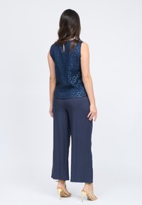 Top senza maniche di un blu scuro con un motivo esagonale testurizzato; abbinato a pantaloni a palazzo blu e sandali con tacco alto dorati. Vista posteriore.