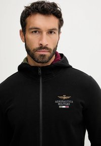 Felpa nera con zip e cappuccio, con logo ricamato "Aeronautica Militare" e dettagli con bandiera italiana sul petto. Materiale in misto cotone.