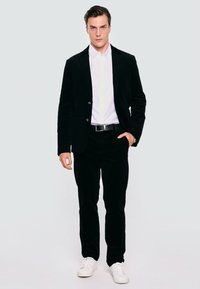 Czarny garnitur z korduroy z dopasowaną marynarką, biała koszula z guzikami, czarny pasek i casualowe białe sneakersy. Gładka faktura i dopasowany krój.