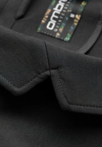 Primer plano de un cuello de prenda de tela negra con costuras precisas y una etiqueta de marca colorida parcialmente visible.