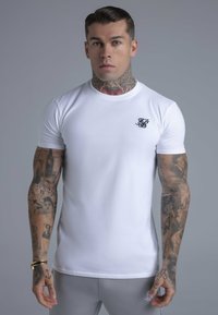 Hvid tætsiddende t-shirt lavet af bomuld, med en lille sort logo på brystet, glat tekstur og korte ærmer. Tatoveringer synlige på armene.