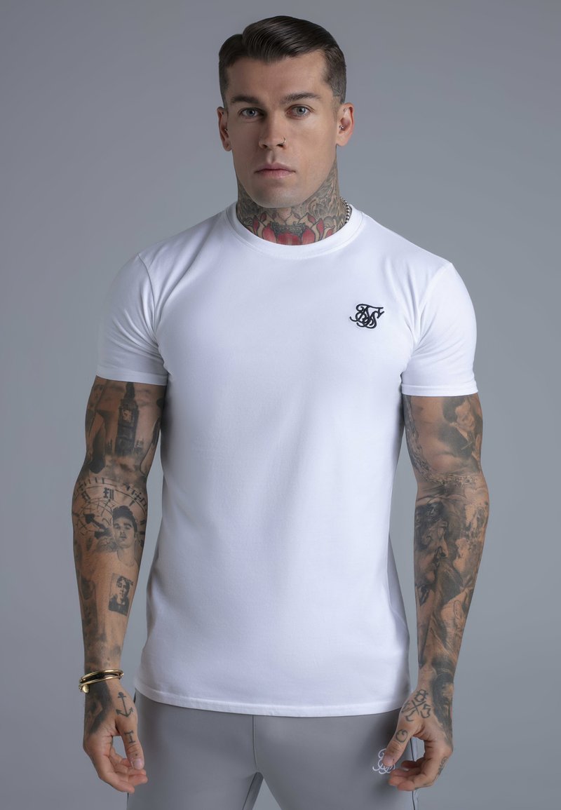 Hvid tætsiddende t-shirt lavet af bomuld, med en lille sort logo på brystet, glat tekstur og korte ærmer. Tatoveringer synlige på armene.