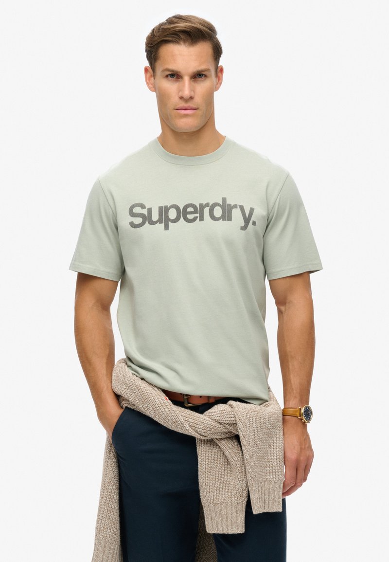 Camiseta de algodón verde claro con gráfica de "Superdry" en gris oscuro, con cuello redondo. Suéter marrón atado a la cintura, pantalones azul marino, reloj de pulsera visible.