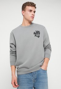 Szürke pulóver, bordázott mandzsettával és aljjal, fekete logóval a bal mellen. Világoskék farmerrel viselve.