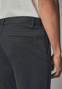 Dunkelgraue Hose mit schlankem Design, aus glattem Stoff gefertigt. Verfügt über eine Gesäßtasche mit horizontaler Öffnung und minimaler Naht.