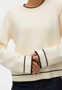 Krēmīgas krāsas džemperis ar melnām akcentētām līnijām aprocēs un apakšmalā, ar mīkstu tekstūru un apaļu apkakli, stilizēts kopā ar tumšām džinsām.