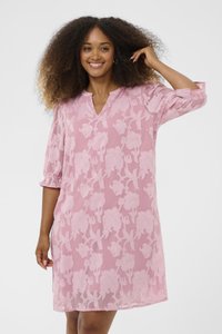 Leende kvinna med lockigt hår som bär en rosa blommig spetsklänning med 3/4-ärmar, stående mot en enkel vit bakgrund.