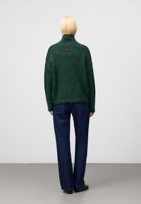 Ciemnozielony sweter z dzianiny z wysokim kołnierzem i teksturowanym wzorem, w połączeniu z ciemnoniebieskimi spodniami typu straight-leg i czarnymi butami.